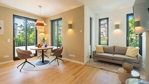Ein modernes Wohnzimmer mit einem runden Esstisch, zwei Stühlen, einer Couch und einem Couchtisch. Große Fenster sorgen für natürliches Licht und Ausblick auf die grüne Umgebung.