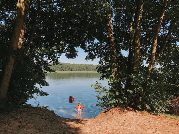 Ein See, umgeben von Bäumen, mit einer Person im Wasser und einer am Ufer.