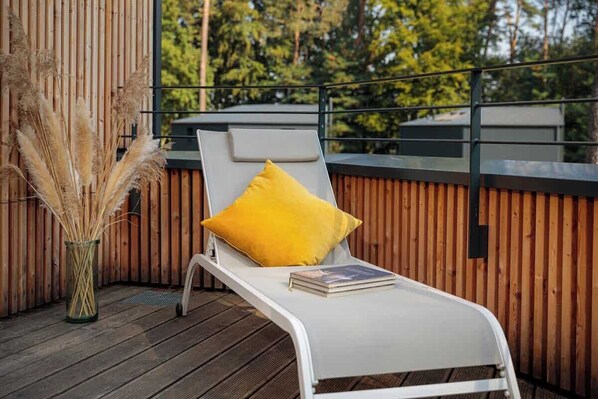 A sun lounger with a yellow pillow and a book on a wooden deck with a wooden wall and a metal railing in the background. - Haus 1 - ELF AM SEE (Templin)