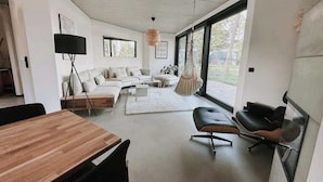 A modern living room with a wooden dining table, a sofa, a hanging chair, and a large window providing natural light. - Haus 1 - ELF AM SEE (Templin)