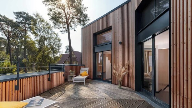 A modern outdoor deck with wooden flooring, a white lounge chair with a yellow cushion, and a glass door leading to an interior space. - Haus 1 - ELF AM SEE (Templin)