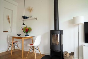 Een moderne woonkamer met een zwarte houtkachel, een eettafel met stoelen, een televisie en een lamp.