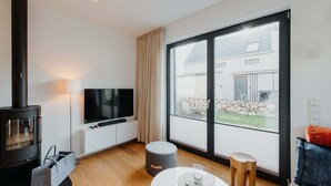 A modern living room with a wood-burning stove, a flat-screen TV, a large sliding glass door to the outdoors, and a cozy seating area. - Holiday Home SandDorn 1 (Pruchten)