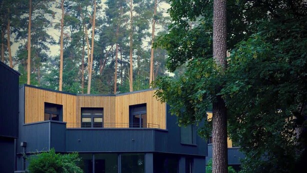 A modern building with a wooden facade, surrounded by tall trees and greenery. - Elf am See - House 7 (Templin)