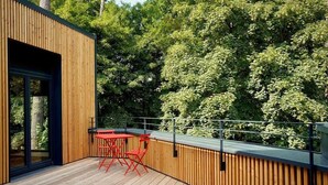 A wooden deck with a table and chairs, surrounded by dense green trees. - Elf am See - House 7 (Templin)