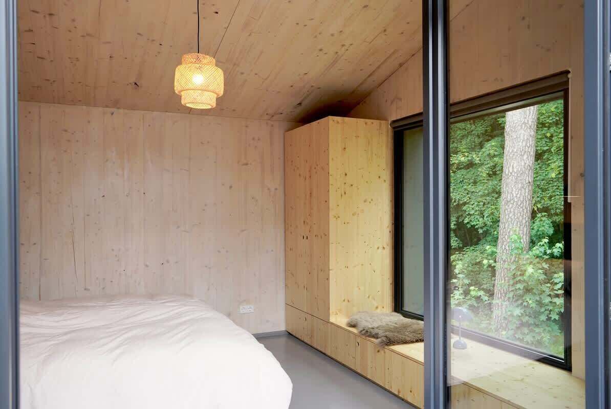 Quarto moderno com teto de madeira, uma janela ampla com vista para árvores e uma cama com colchão branco.