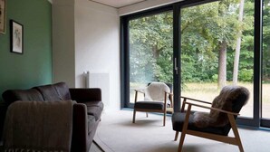 A modern living room with a large window, a sofa, a chair, and a radiator. - Elf am See - House 7 (Templin)