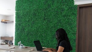 Una mujer sentada a una mesa con una computadora portátil en una habitación con pared verde y sillas blancas.