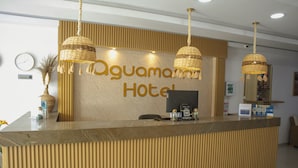 Un mostrador de recepción con un letrero que dice 'Oguamama Hotel', un reloj de pared y una barra de recepción con una computadora y algunos objetos encima.