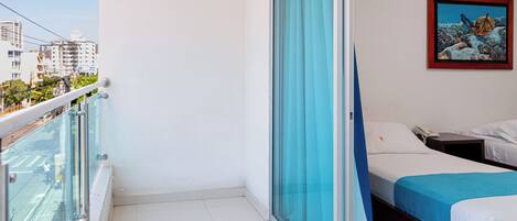 Habitación de hotel con balcón, cama con ropa blanca, un cuadro en la pared y vista panorámica de la ciudad.