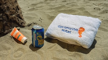A towel with "Aquamarina Hotel" embroidered on it, a can of "Coca-Cola," and a tube of sunscreen on a sandy surface.