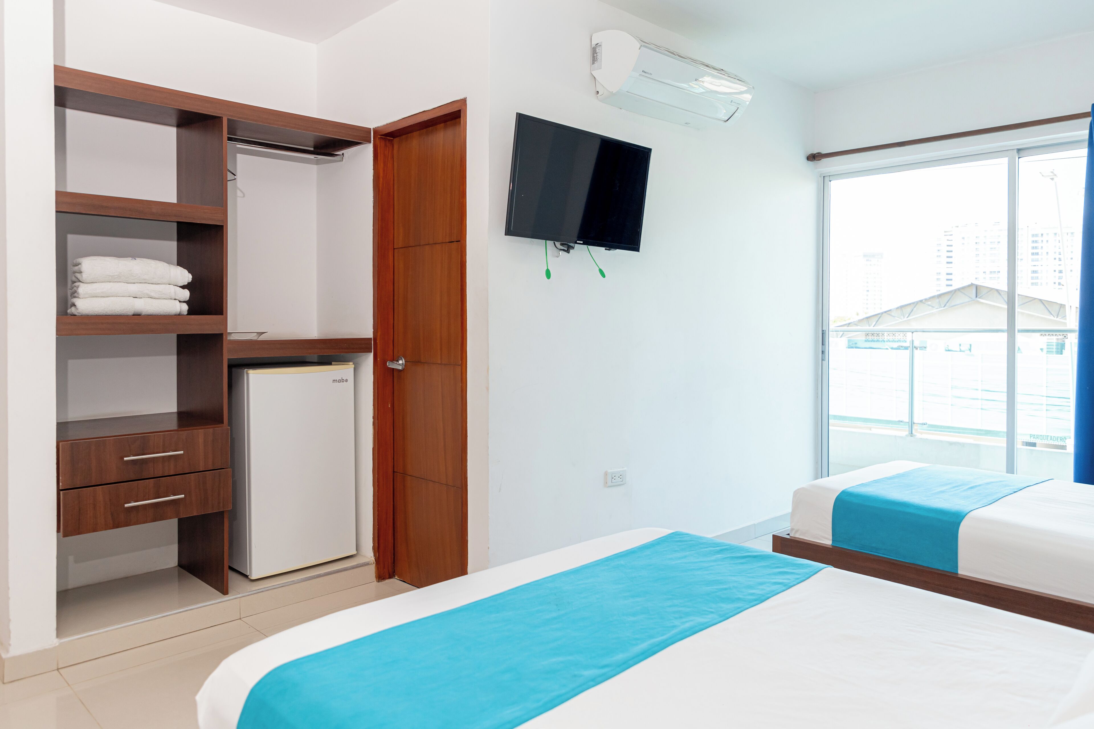 A hotel room with a wardrobe, a television, a bed, and a window with a view of buildings.