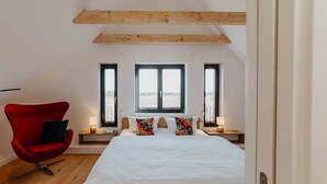 A bedroom with a large bed, two bedside tables, a red chair, and a window with a view. - Holiday Home SandDorn 2 (Pruchten)