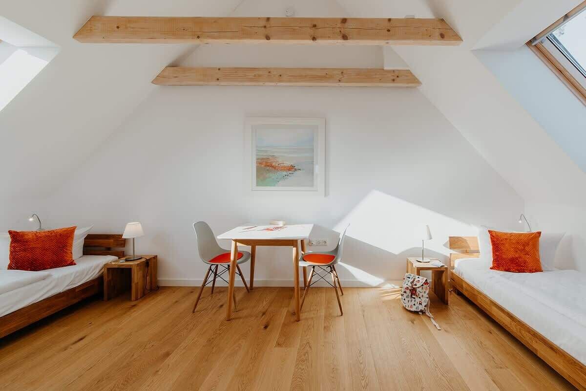 A modern loft bedroom with a sloped ceiling, wooden beams, a dining table with chairs, and a bed with red pillows.