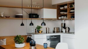 A modern kitchen with white cabinets, wooden shelves, and a dining area with a wooden table and chairs. - Holiday Home SandDorn 2 (Pruchten)