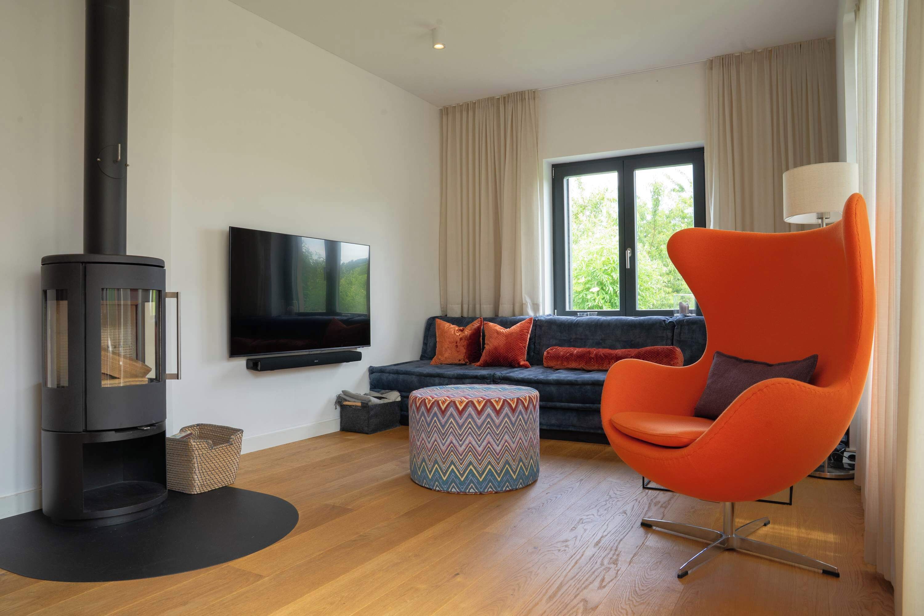 Un salon moderne avec un poêle à bois noir, un grand téléviseur à écran plat, un canapé de couleur sombre agrémenté de coussins orange et un fauteuil orange vif.