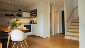 A modern kitchen with white cabinets, wooden flooring, and a dining area with a white chair and a vase of flowers. - Holiday Home SandDorn 2 (Pruchten)