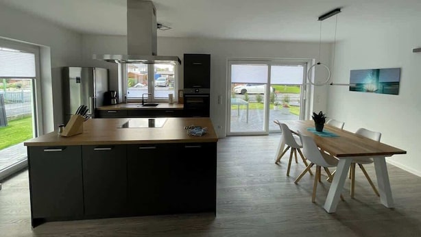 Una cocina moderna con una isla central, un comedor con mesa y sillas de madera, y grandes ventanales que ofrecen vistas al exterior.