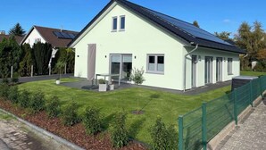 Una casa moderna de dos pisos con techo inclinado, paneles solares, césped bien cuidado y cerca verde.