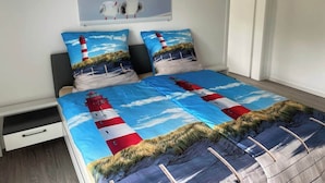 Una cama con una colcha de motivos de faro, una mesita de noche pequeña y un cuadro en la pared con gaviotas.