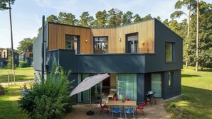 Et moderne hus i to etager med fladt tag, store vinduer og en træterrasse med udendørs møbler.