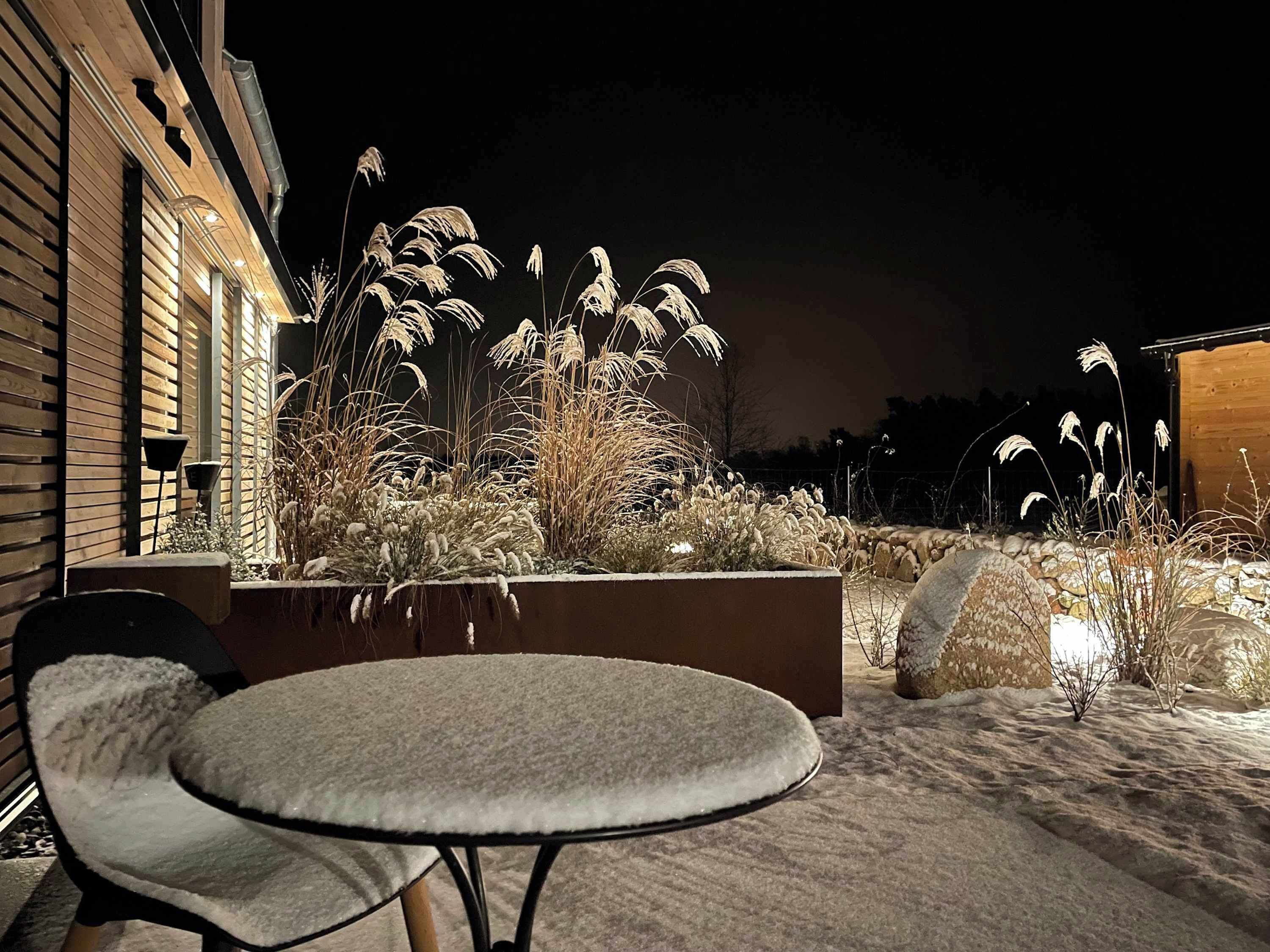 Een terras met een ronde tafel en stoelen, omringd door hoge grassen en een houten gebouw op de achtergrond.