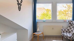 Ett sovrum med en säng, en stol, ett litet bord med en modell av ett skepp, ett element och ett fönster med gardiner.