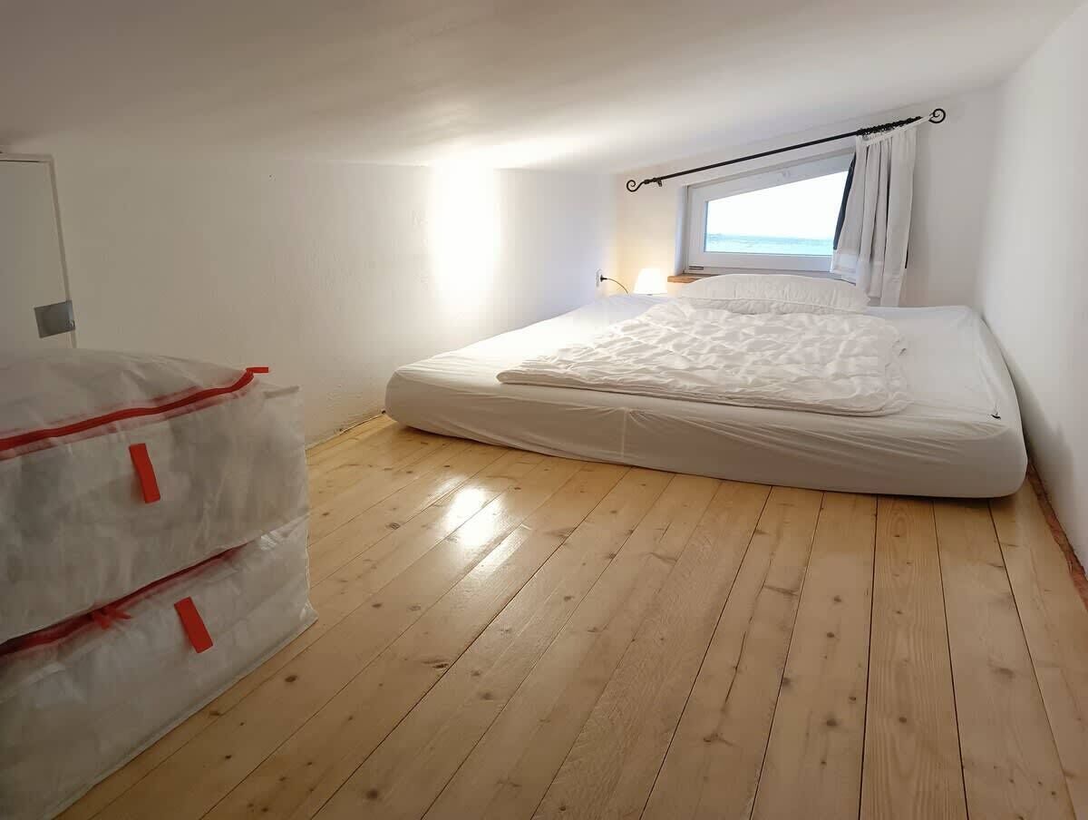 Ein Zimmer mit Holzboden, einem Bett mit weißer Bettwäsche, einem Fenster mit Vorhängen und einer an der Wand befestigten Leuchte.