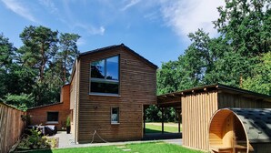 A modern wooden house with a large glass window, a covered outdoor seating area, and a wooden barrel structure.