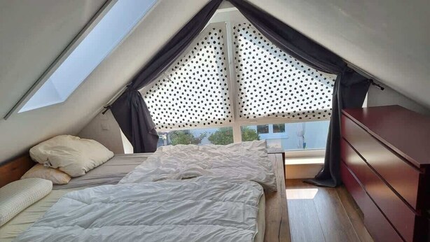 Ein kleines, dreieckiges Schlafzimmer im Dachgeschoss mit Bett, Holzboden und Blick durch ein Fenster mit Lasterdekor.