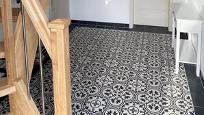 A staircase with wooden railings and a black metal handrail, leading to a room with patterned floor tiles, a white door, and a small table. - Paula (Marlow)
