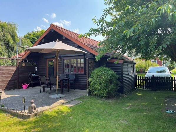 A wooden cabin with a patio, a small shed, and a car parked in the background.