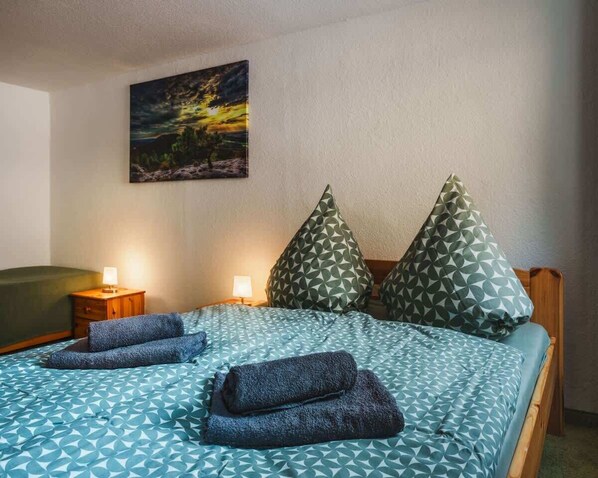 Una cama bien hecha con ropa de cama estampada y dos almohadas, una mesita de noche con lámpara, y un cuadro en la pared.