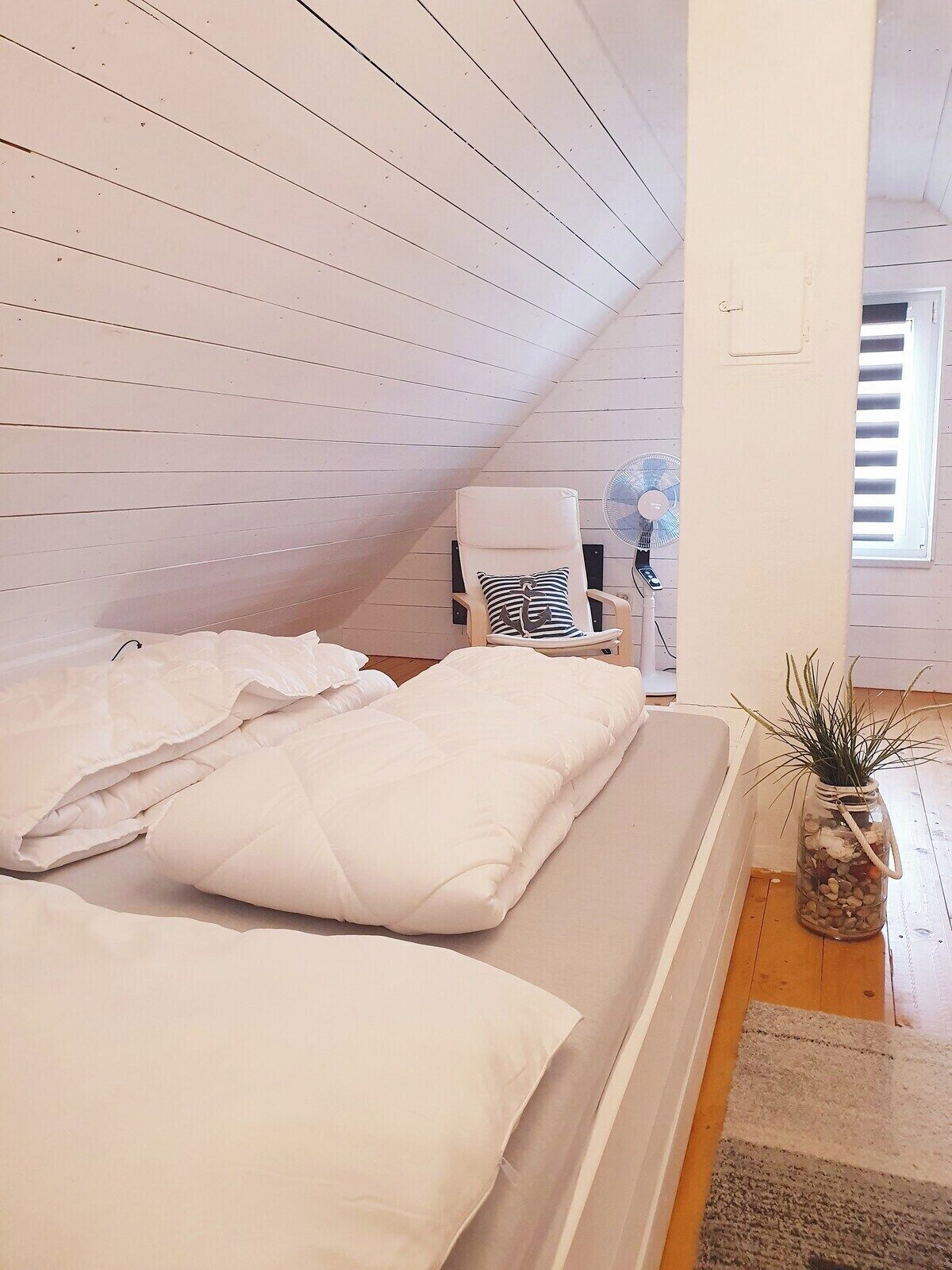 A bedroom with a sloped ceiling, a bed with white bedding, a chair, a fan, and a window with blinds.