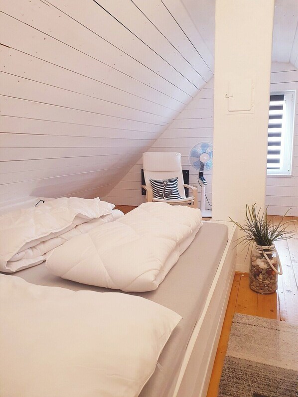 A bedroom with a sloped ceiling, a bed with white bedding, a chair, a fan, and a window with blinds.