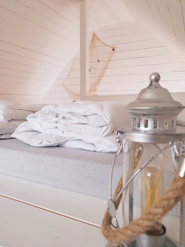 A neatly made bed with folded white linens, a metal lantern with a rope handle, and a white wall with a visible electrical outlet.