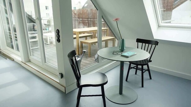 A room with a round table, two chairs, a vase with a flower, and a view of an outdoor deck with a wooden table and chairs. - DUS-Loft (Düsseldorf)
