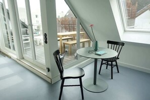 Een kamer met een ronde tafel, twee stoelen, een vaas met een bloem en uitzicht op een terras buiten met een houten tafel en stoelen.