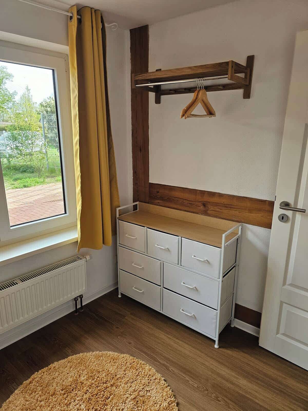 Habitació amb finestra, un radiador, una petita còmoda blanca amb calaixos, una prestatgeria de fusta i una porta.