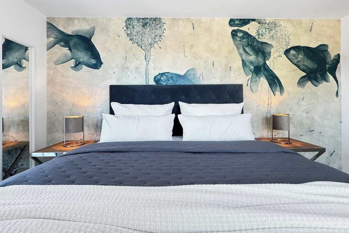 Ein Bett mit dunklem Kopfteil, weißen Kissen und einer gesteppten Decke. Zwei Nachttische mit Lampen und ein großes Wandbild von Fischen sind ebenfalls zu sehen.
