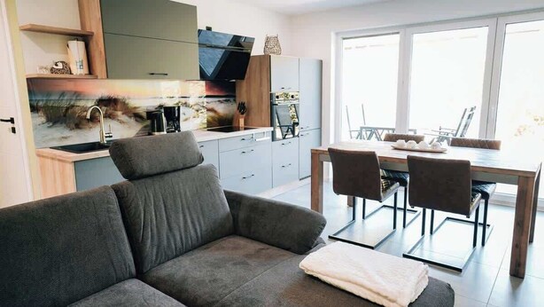 Una sala moderna con un sofá gris, un comedor con mesa y sillas de madera, y una cocina con armarios grises y un ventanal grande.