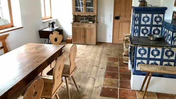 Een rustieke keuken met een houten tafel en stoelen, een kast met glazen deuren en een betegelde kachel.