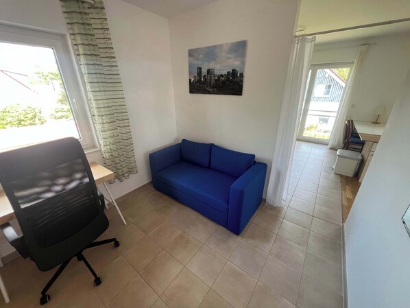 Une pièce avec un canapé bleu, un fauteuil de bureau noir, un tableau représentant un paysage urbain accroché au mur et une fenêtre avec des rideaux.