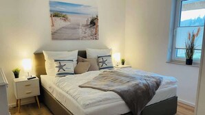 Un dormitorio con cama, mesitas de noche, una ventana con vistas y un cuadro con temática de playa.