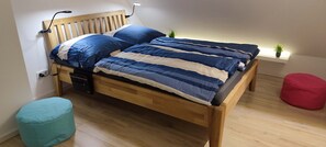 A bed with a wooden headboard, a bedside table with a lamp, a small plant on a shelf, and a red ottoman.