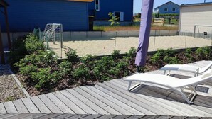 Eine Holzterrasse mit zwei weißen Liegestühlen, ein Garten mit Sträuchern und Blumen, und ein Fußballtor im Hintergrund.