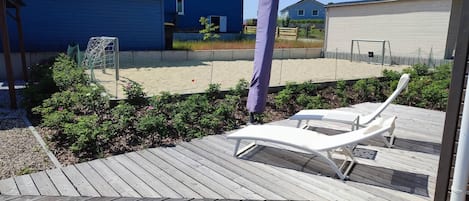 Une terrasse en bois avec deux chaises longues blanches, un jardin agrémenté d’arbustes et de fleurs, et un but de football en arrière-plan.