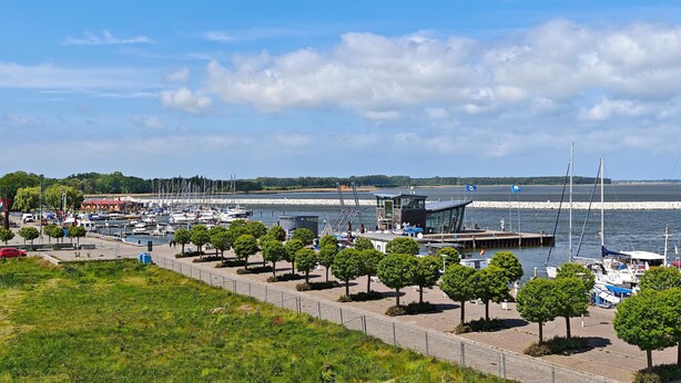 A marina with boats docked, a pedestrian walkway lined with trees, and a view of the water and distant shoreline. - Barther  Hafensonne (Barth)