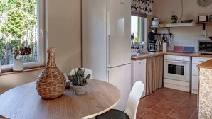 Una cocina con una mesa redonda de madera, sillas blancas, un refrigerador, una estufa y una ventana con vistas a árboles.
