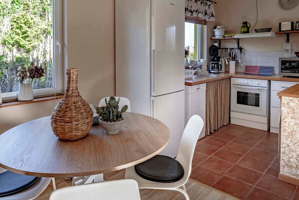 A kitchen with a round wooden table, white chairs, a refrigerator, a stove, and a window with a view of trees.